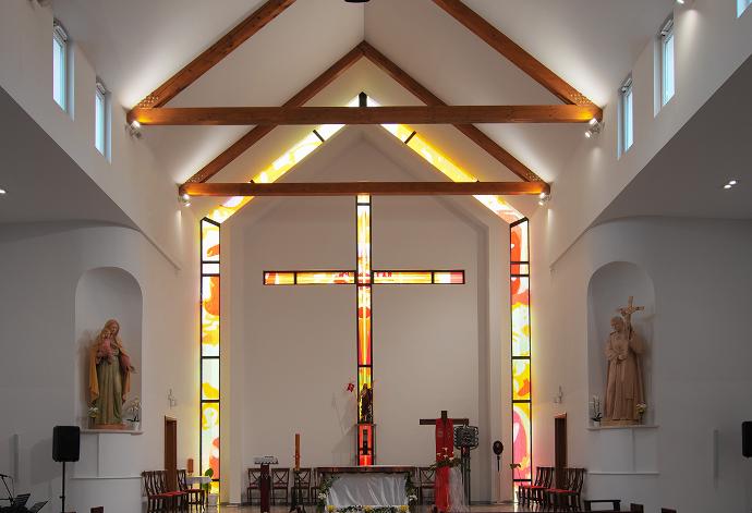 Unutrašnjost suvremene crkve s drvenim gredama, statuama svetaca i vitrajem iza oltara